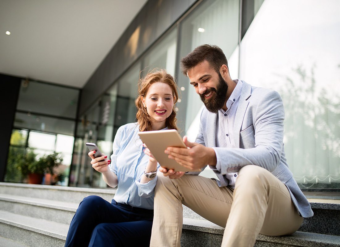 Contact - Portrait of a Smiling Young Business Woman and Man Using Technology While Sitting Outside a Modern Glass Office Building During a Lunch Break